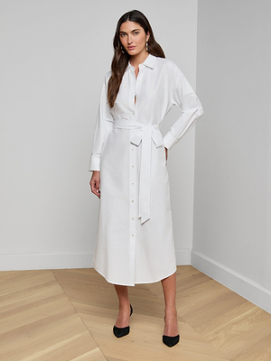 A woman stands indoors against a plain background wearing the Ridley Shirt Dress—an organic cotton white shirt dress with long sleeves and a tied waist belt. She pairs it with black heels and poses confidently, one hand on her hip, showcasing her long dark hair.