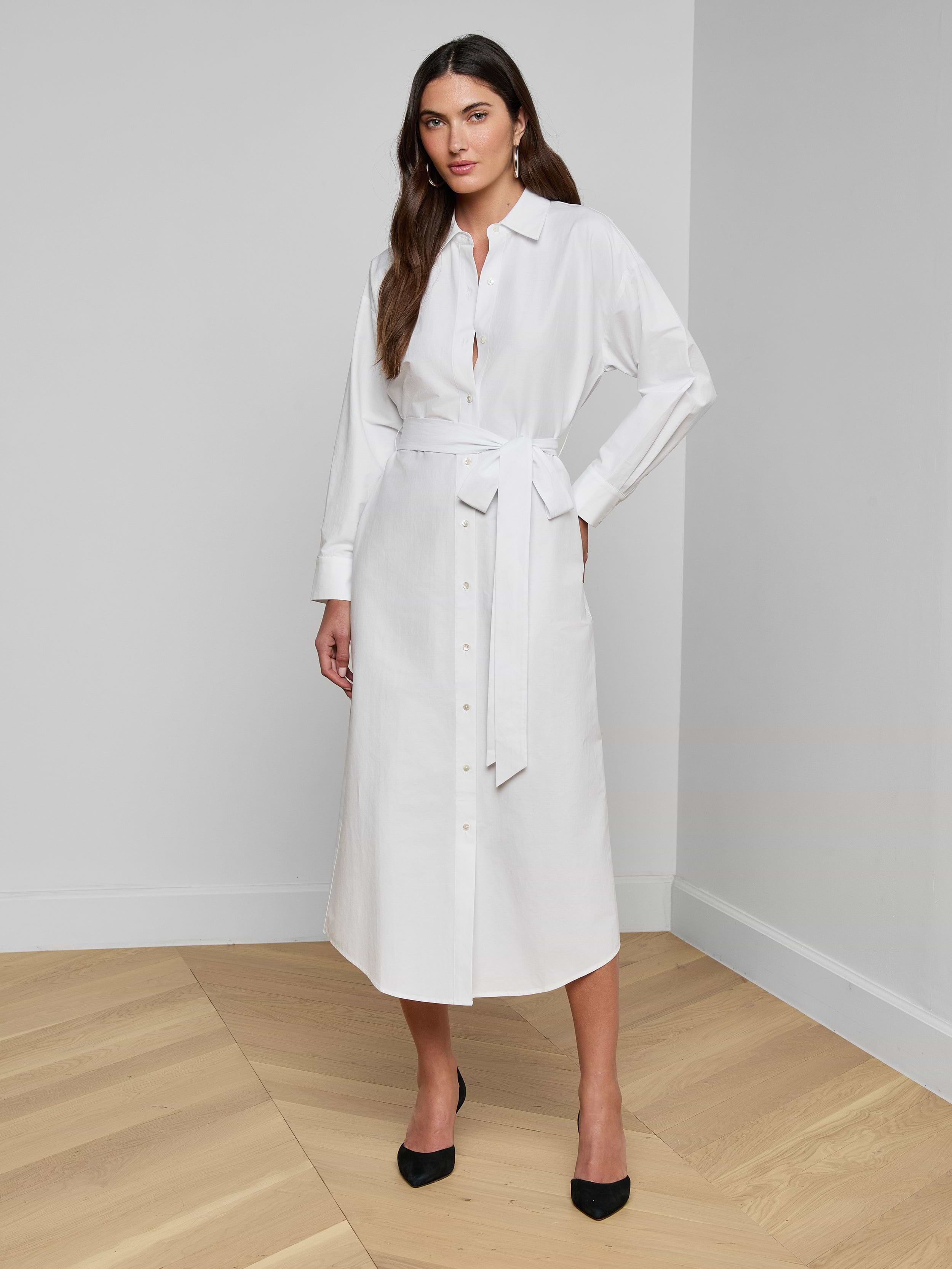 The woman is wearing the Ridley Shirt Dress, a long-sleeved, oversized white dress crafted from organic cotton, complete with a matching waist tie. She stands against a plain white background with one hand on her hip and pairs the look with black heels.