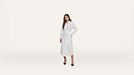 A woman stands against a plain white background, wearing a long-sleeved, belted white dress with black high heels.
