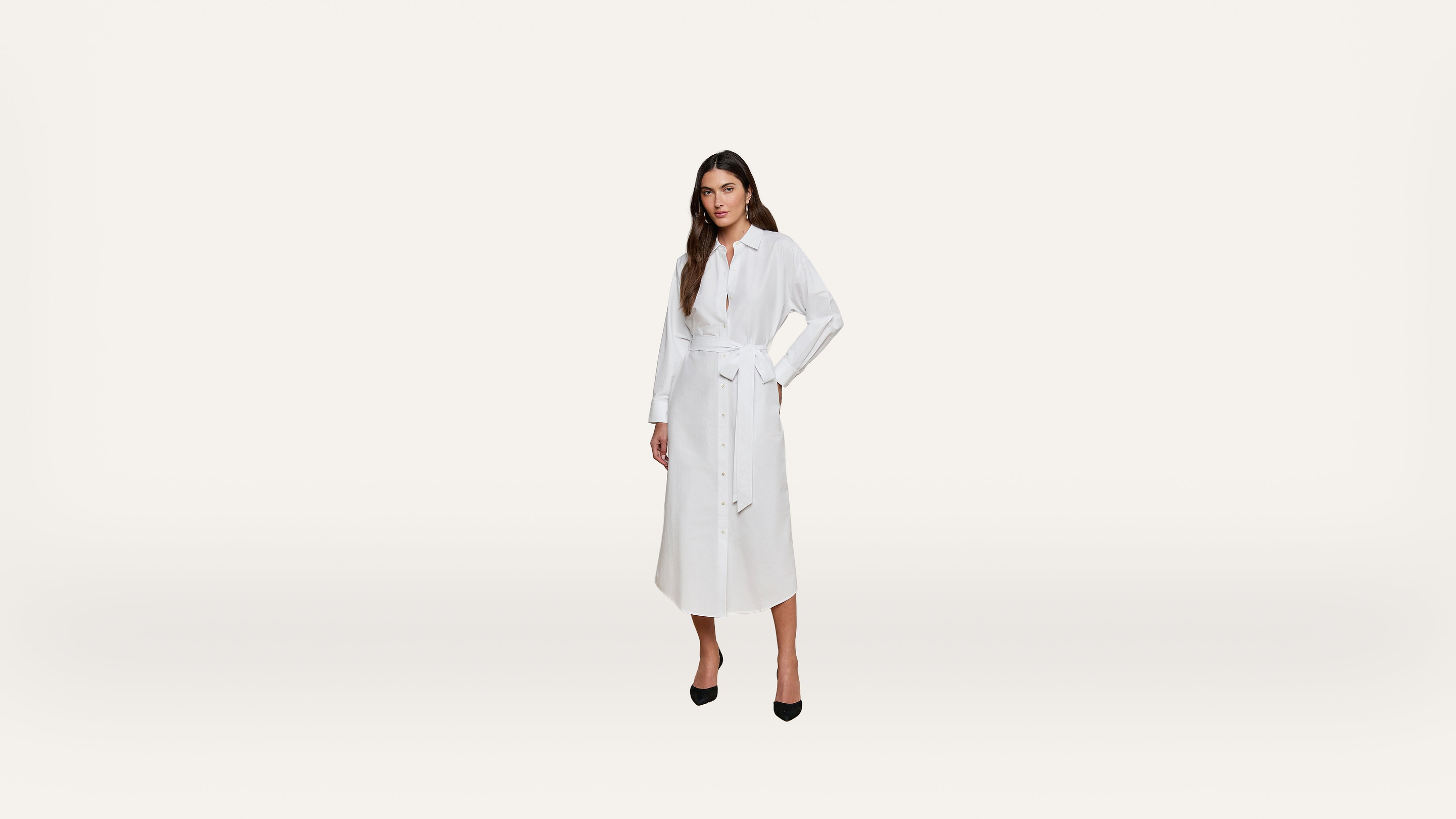 A woman stands against a plain white background, wearing a long-sleeved, belted white dress with black high heels.