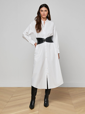A woman poses confidently indoors wearing the Ridley Shirt Dress in organic cotton white, styled with a wide black textured belt and black knee-high boots. She has long brown hair and stands against a plain background.