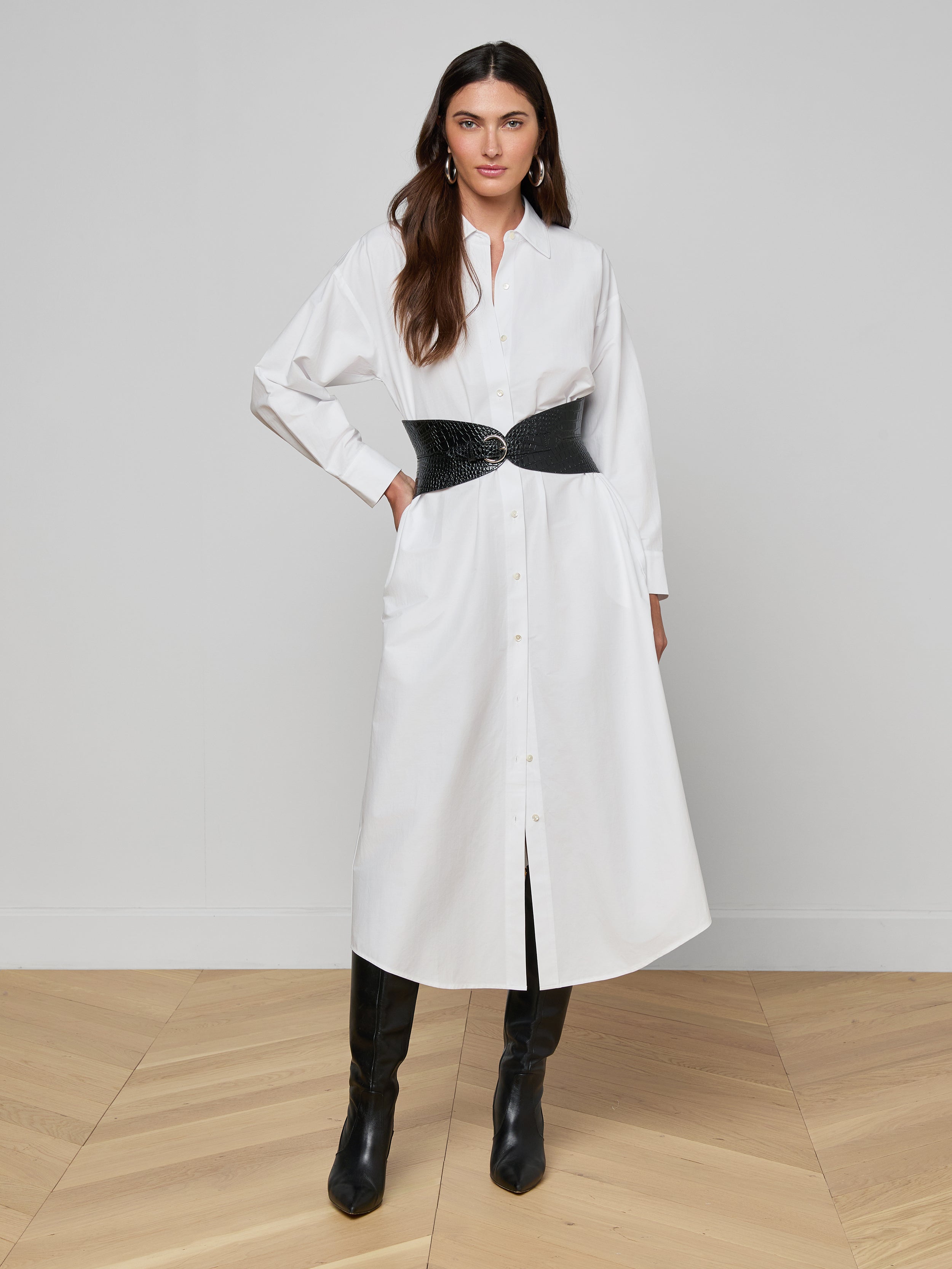 A woman stands against a plain white background, wearing the Ridley Shirt Dress—an oversized fit white shirt dress made from organic cotton—styled with a black textured belt and black knee-high boots.