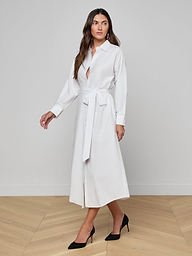 A woman wearing the Ridley Shirt Dress—a long-sleeve, oversized-fit, white shirt dress made from organic cotton and styled with a waist belt—stands against a plain light background in black heels, gazing slightly to her left.