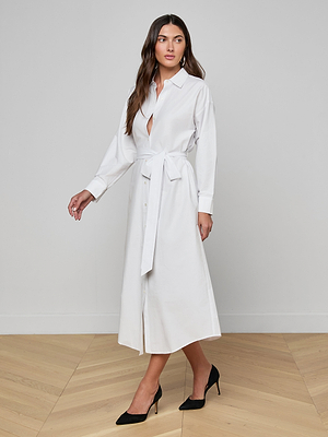 A woman poses indoors in the Ridley Shirt Dress, a long sleeve white organic cotton style with a belted waist and front buttons, paired with black pointed-toe heels on a light wood floor against a plain white wall.