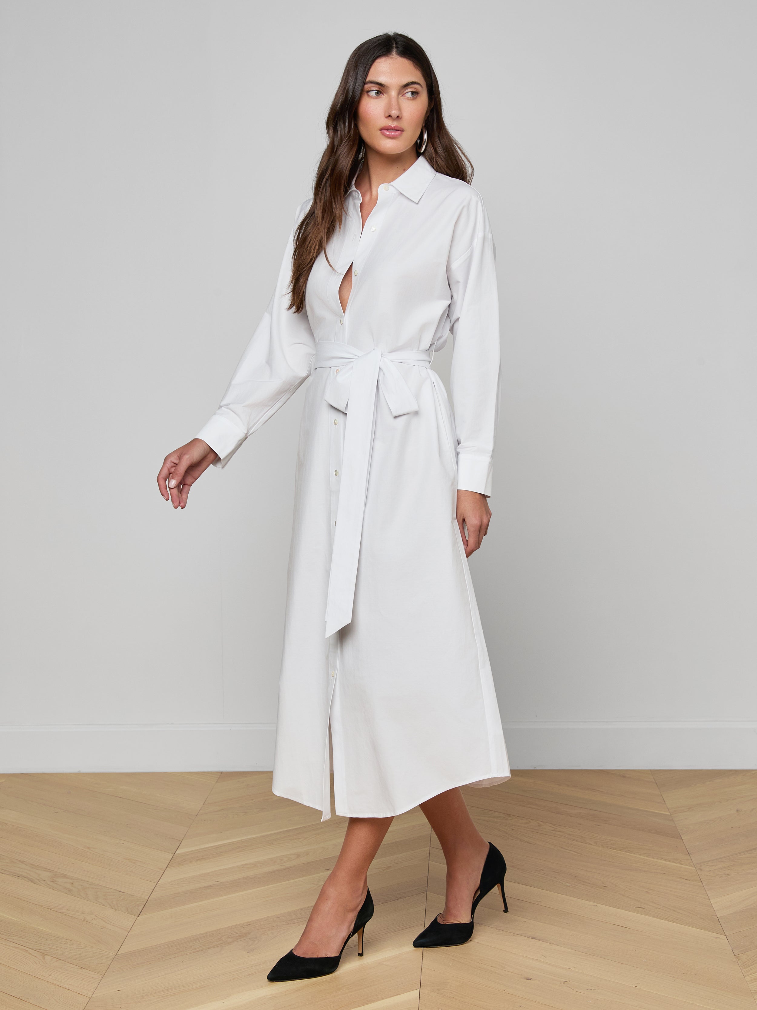 A woman wearing the Ridley Shirt Dress—a long-sleeve, oversized-fit, white shirt dress made from organic cotton and styled with a waist belt—stands against a plain light background in black heels, gazing slightly to her left.