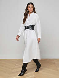 Wearing the Ridley Shirt Dress—an oversized, white, organic cotton shirt dress—a woman stands against a plain white background, styled with a black wide belt and black heeled boots.