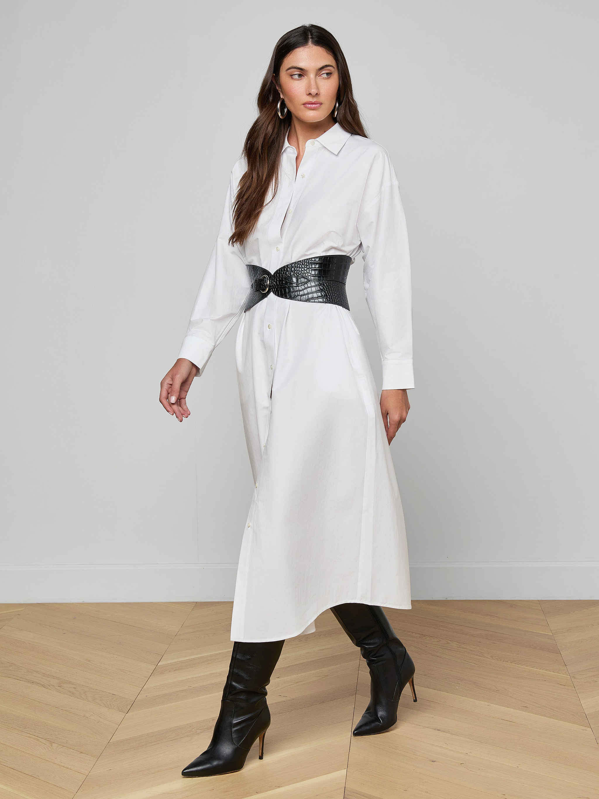 Wearing the Ridley Shirt Dress—an oversized, white, organic cotton shirt dress—a woman stands against a plain white background, styled with a black wide belt and black heeled boots.