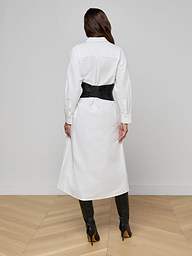 A person stands facing away against a plain white background, wearing the Ridley Shirt Dress in white with an oversized fit, styled with a black wide belt and black knee-high boots.