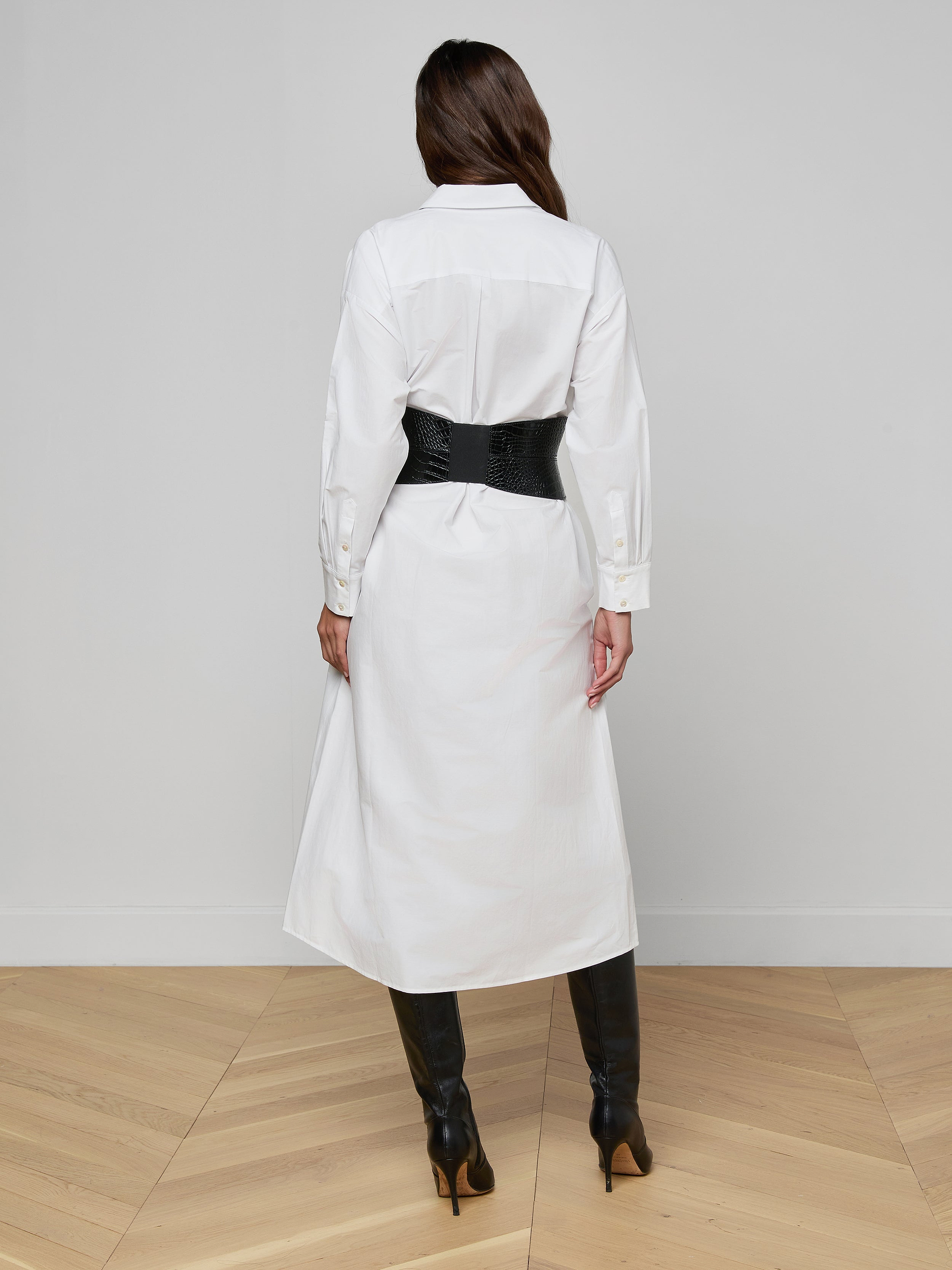 A person stands facing away against a plain white background, wearing the Ridley Shirt Dress in white with an oversized fit, styled with a black wide belt and black knee-high boots.
