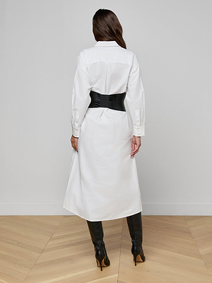 A person with long brown hair, seen from behind, wears the Ridley Shirt Dress with a wide black belt and knee-high black heeled boots, standing on a light wooden floor against a plain wall.