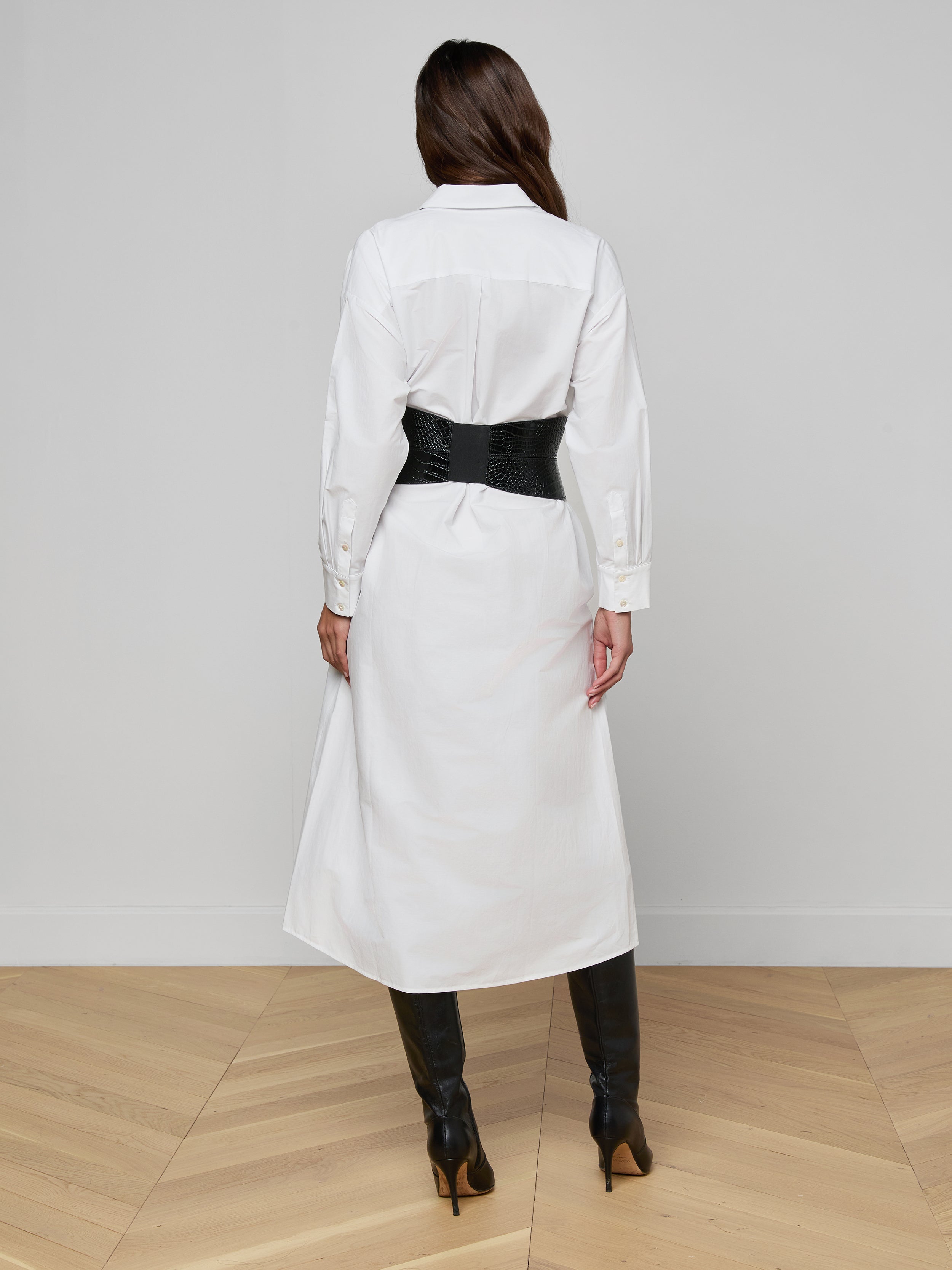 A person stands facing away against a plain white background, wearing the Ridley Shirt Dress in white with an oversized fit, styled with a black wide belt and black knee-high boots.