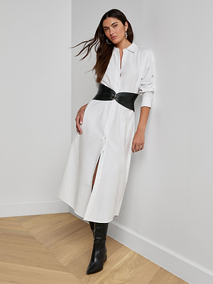A woman stands against a white wall wearing the Ridley Shirt Dress in organic cotton, styled with a wide black textured belt and knee-high black boots. Her dark hair is loose, and she poses with one leg slightly bent.