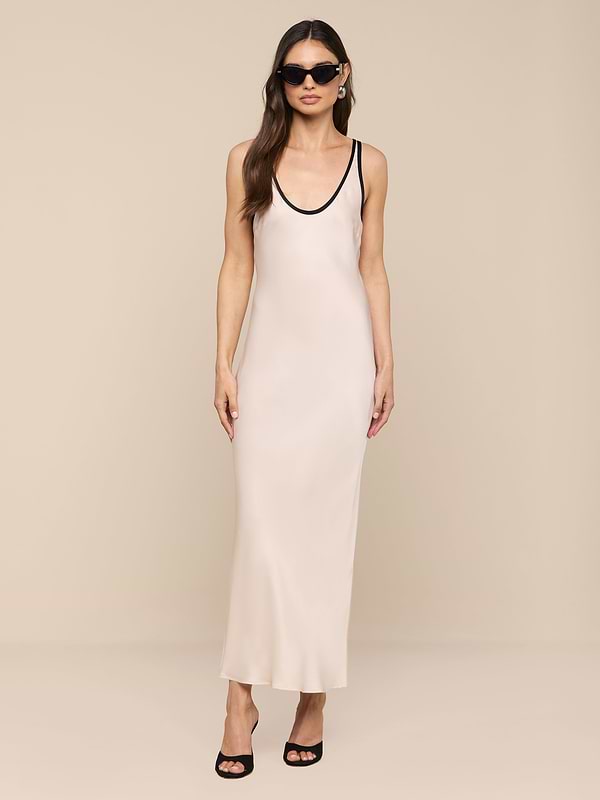 Una donna modella lAkiya Satin Maxi Dress - un abito avorio senza maniche, lungo fino alla caviglia, tagliato in sbieco con finiture nere - abbinato a occhiali da sole, orecchini e tacchi neri aperti su uno sfondo beige.