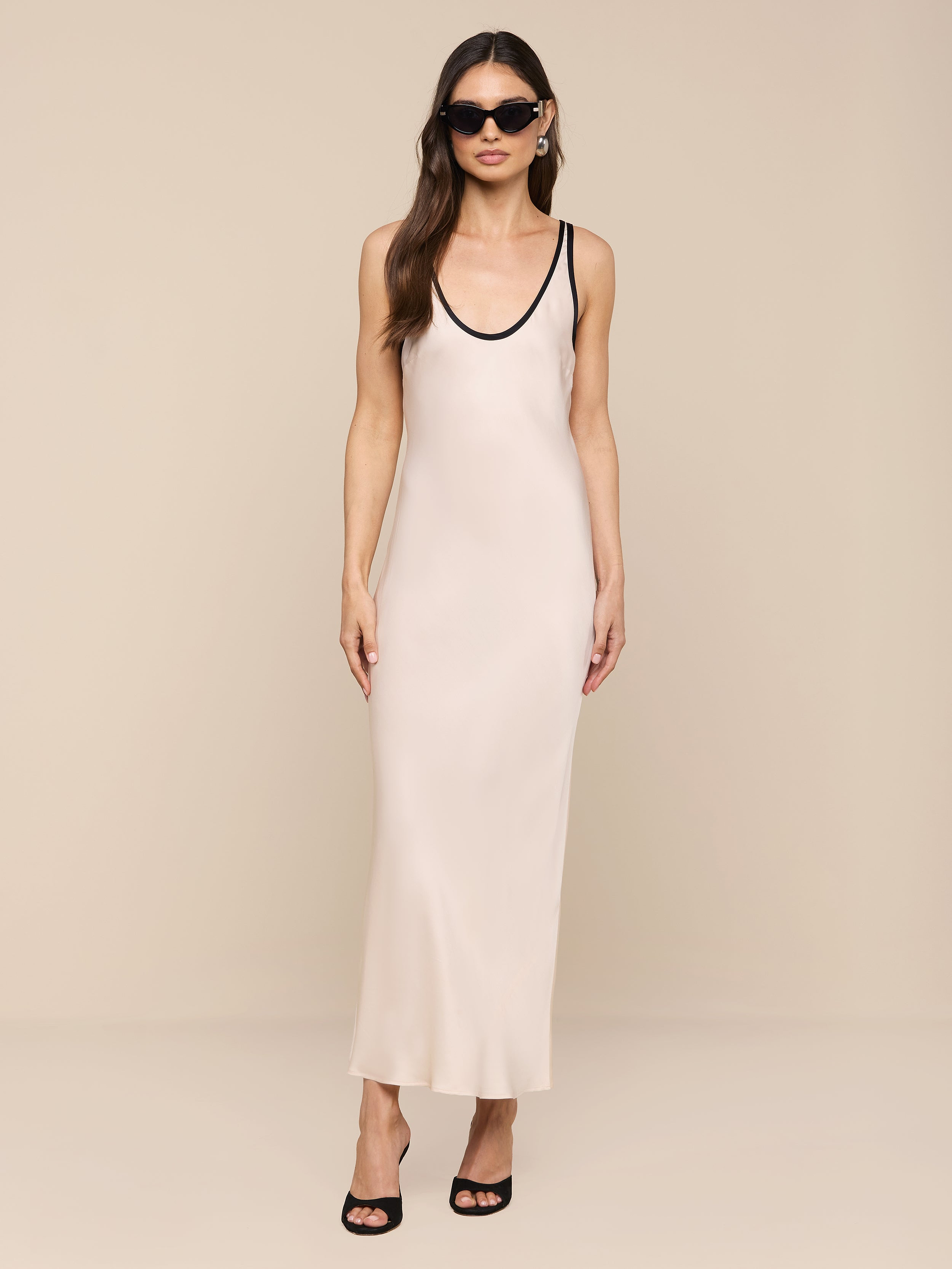 Una donna modella lAkiya Satin Maxi Dress - un abito avorio senza maniche, lungo fino alla caviglia, tagliato in sbieco con finiture nere - abbinato a occhiali da sole, orecchini e tacchi neri aperti su uno sfondo beige.