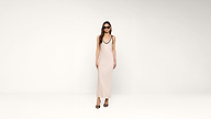 A woman in sunglasses and a long, pale pink sleeveless dress stands against a plain white background, wearing black high-heeled sandals.