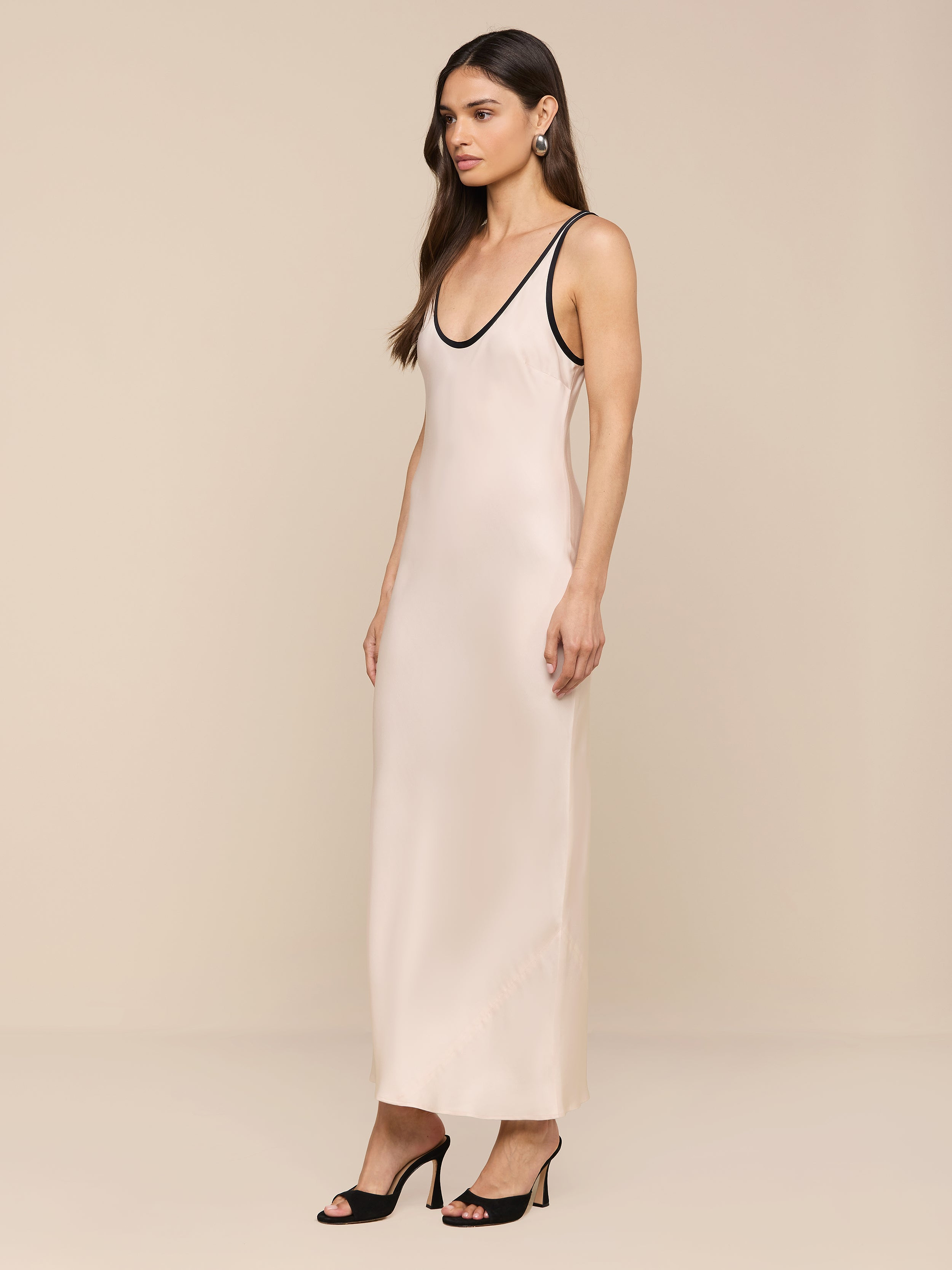 Una donna con lunghi capelli castani ondulati e orecchini di perle si trova in uno studio beige e indossa lAkiya Satin Maxi Dress - senza maniche, tagliato in sbieco con finiture nere - abbinato a tacchi neri open-toe.
