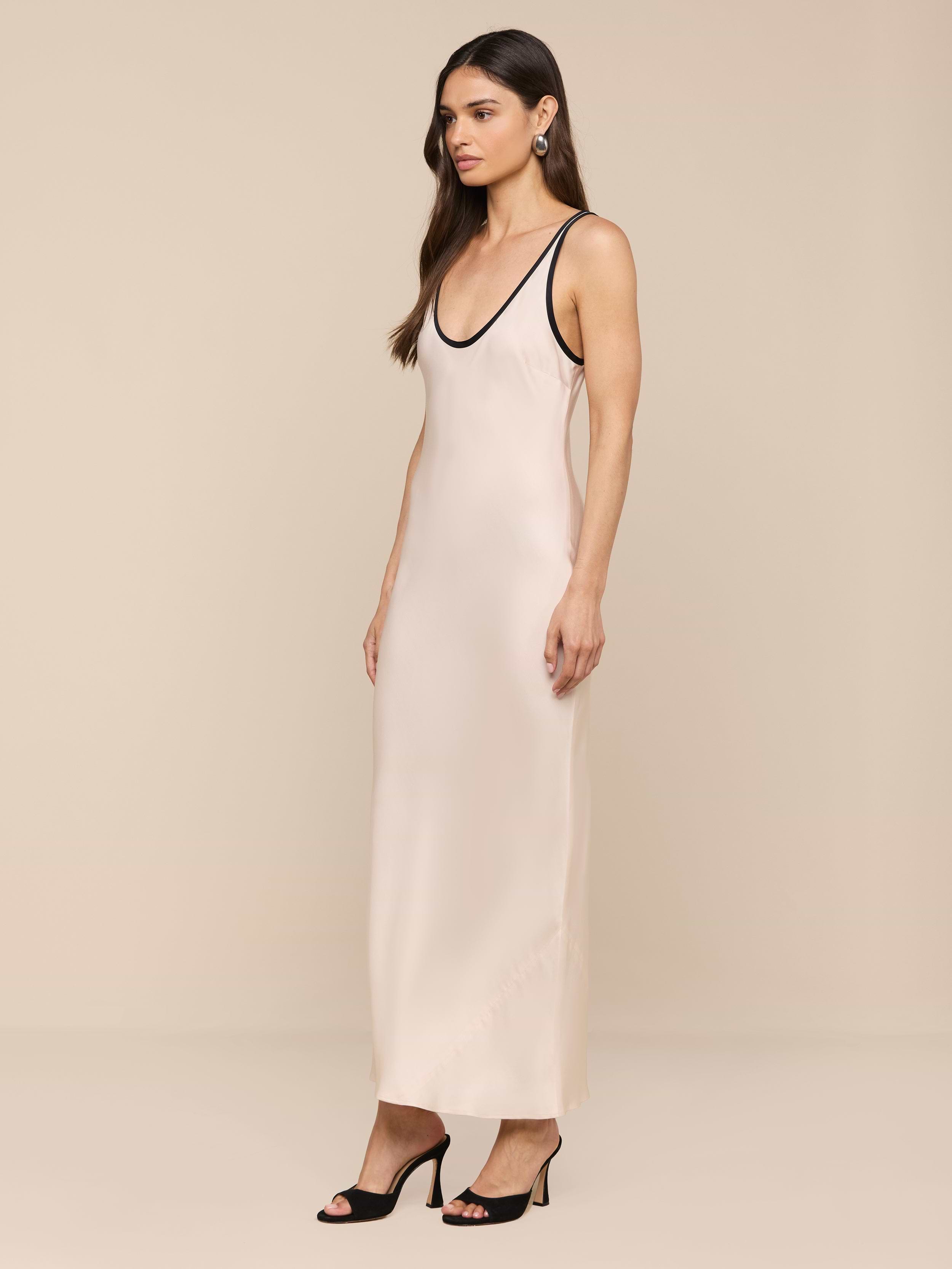 Una donna con lunghi capelli castani ondulati e orecchini di perle si trova in uno studio beige e indossa lAkiya Satin Maxi Dress - senza maniche, tagliato in sbieco con finiture nere - abbinato a tacchi neri open-toe.