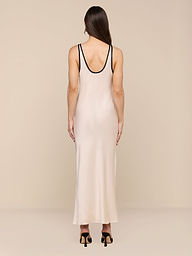 Una donna con lunghi capelli castani indossa il maxi abito Akiya in raso con rifiniture nere e sandali neri con tacco alto su uno sfondo beige: un look chic perfetto da indossare da giorno a sera.