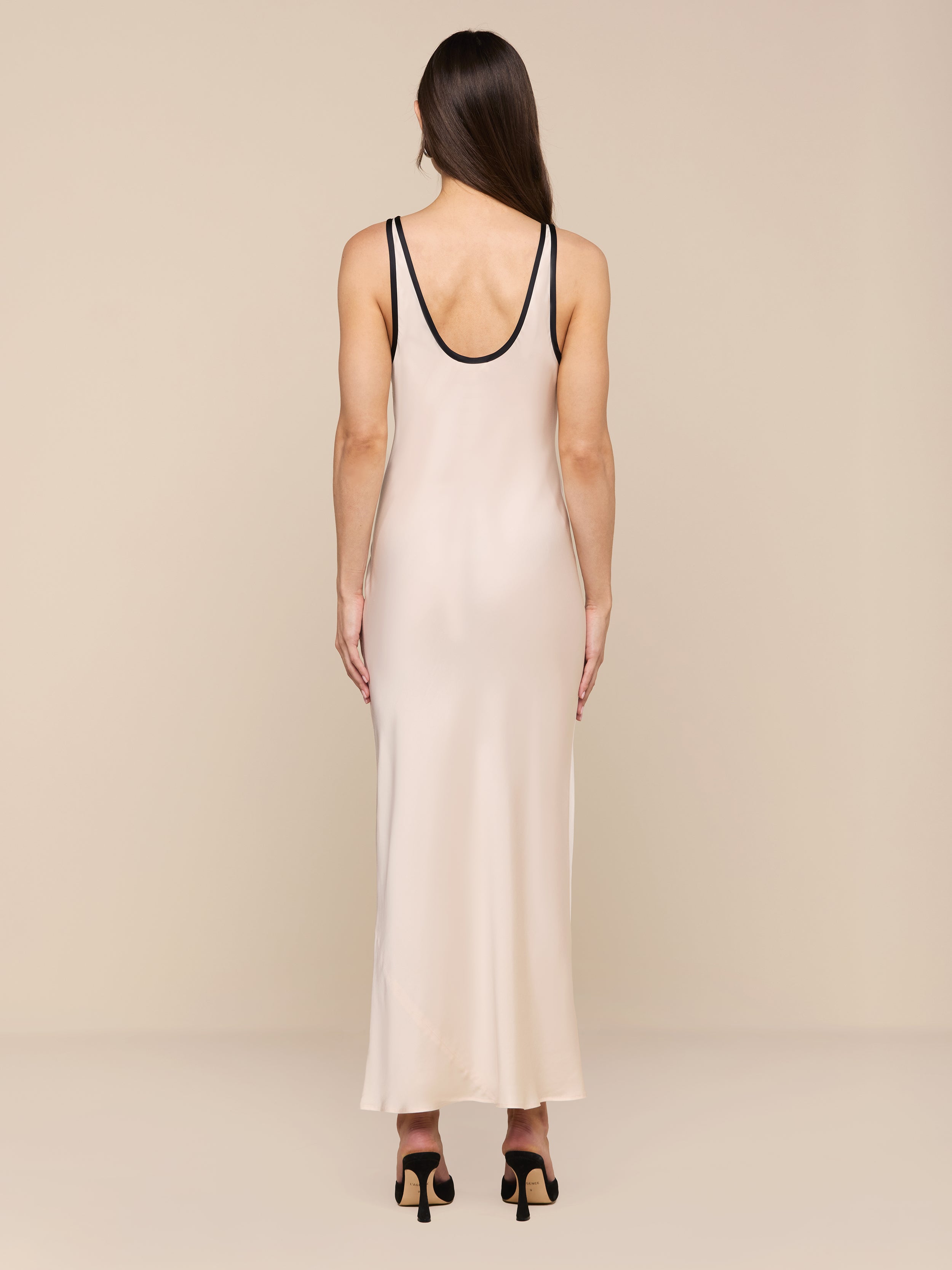 Una donna con lunghi capelli castani indossa il maxi abito Akiya in raso con rifiniture nere e sandali neri con tacco alto su uno sfondo beige: un look chic perfetto da indossare da giorno a sera.