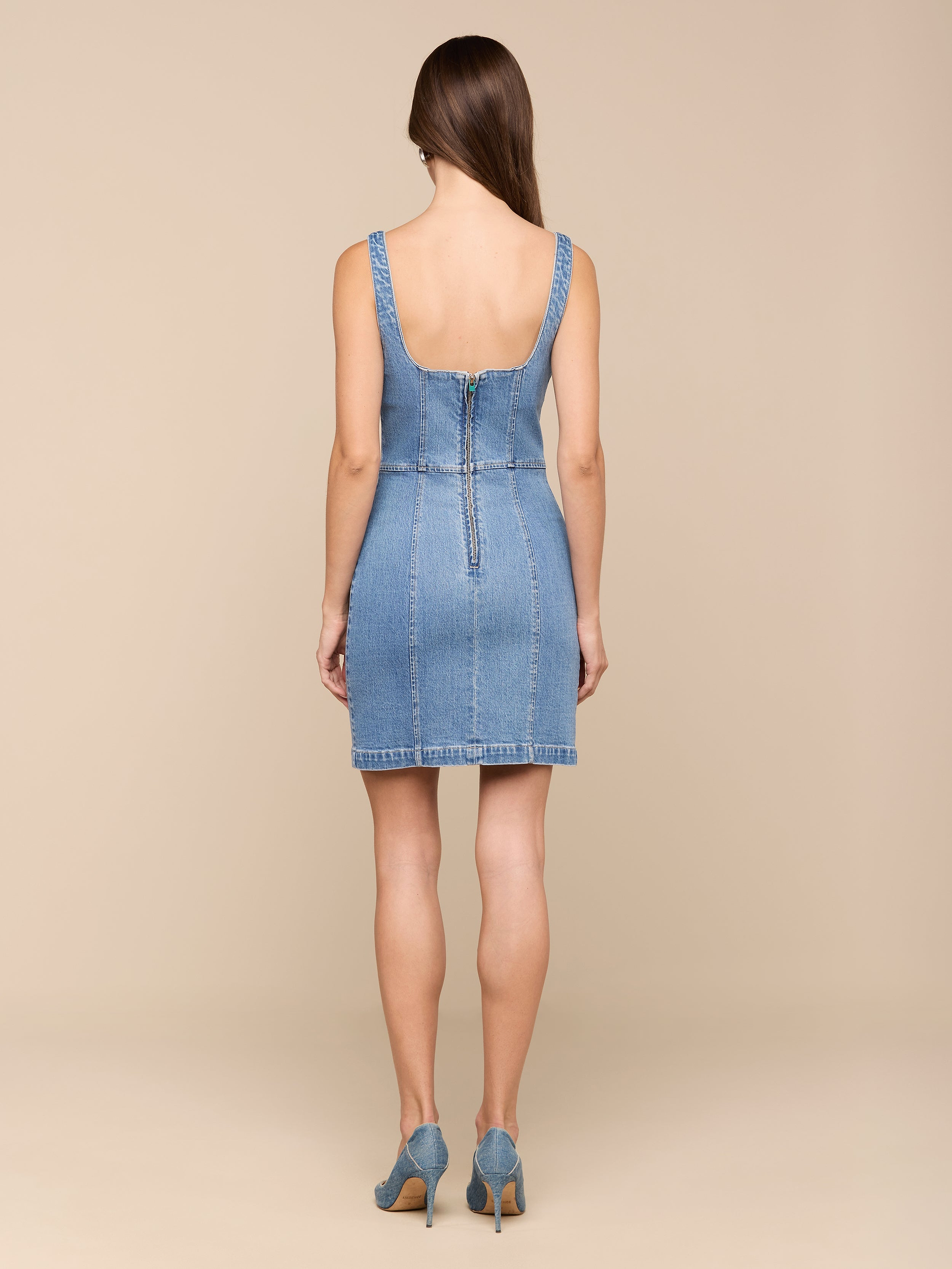 Una donna con lunghi capelli castani è rivolta verso lesterno e indossa il Vynna Dress, un mini abito di LAGENCE in denim elasticizzato blu chiaro con cerniera posteriore, abbinato a tacchi in denim abbinati, su uno sfondo beige semplice.