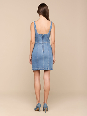 Une femme aux longs cheveux bruns est de face, portant la robe Vynna - une mini robe en denim extensible bleu clair de LAGENCE avec une fermeture éclair dans le dos - associée à des talons en denim assortis, sur un fond beige uni.