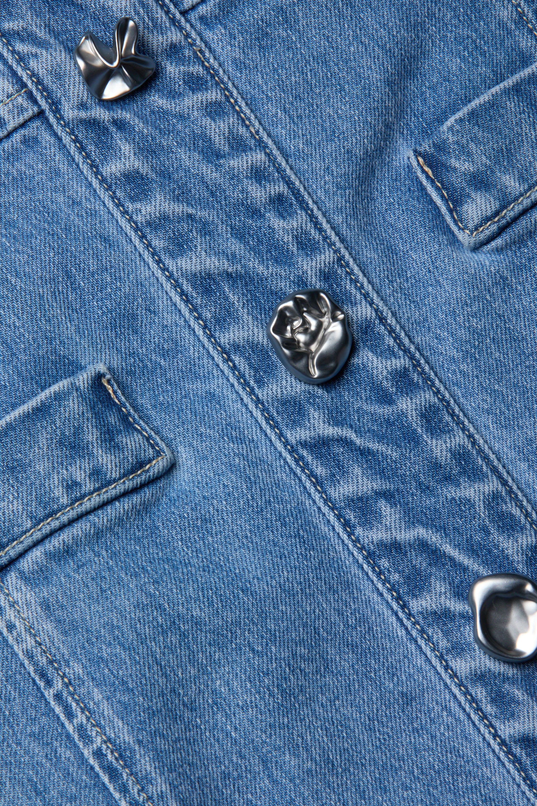 Primo piano del Vynna Dress, un capo in denim blu elasticizzato di LAGENCE, caratterizzato da due tasche sul petto e da bottoni metallici argentati a forma di fiori astratti.