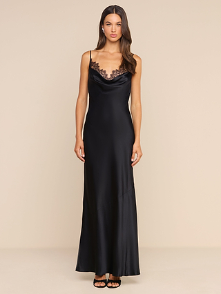 A woman stands in the Selene Silk Gown, a long black Chantilly lace dress with satin trim and thin straps, paired with black heeled sandals against a neutral background.
