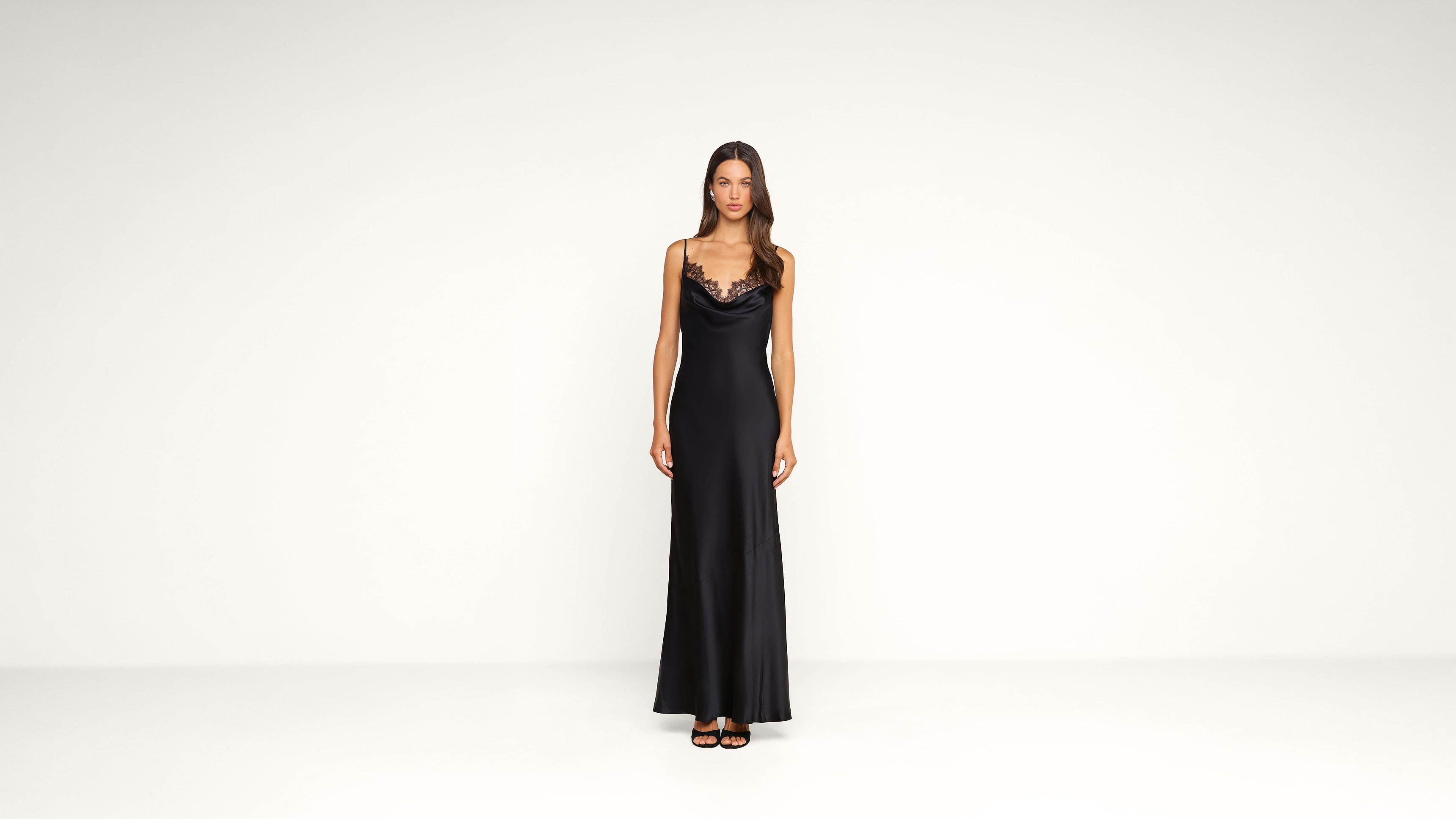 A woman stands against a plain white background, wearing a long black satin dress with thin straps and lace trim. She has straight brown hair and is wearing black heeled sandals.