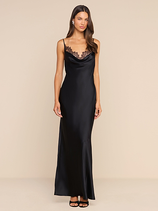 A woman wearing the Selene Silk Gown—a long, black slip dress with lace trim at the neckline and thin straps—stands against a plain beige background. Reminiscent of a classic black silk evening gown, she has long brown hair and is wearing black heels.