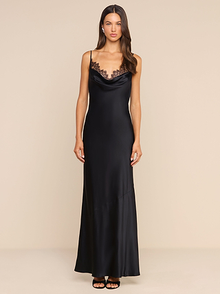 A woman stands against a plain beige background, wearing the Selene Silk Gown—a black silk evening dress featuring thin straps and delicate lace trim at the neckline. The gown flows elegantly to a maxi length, and she completes her look with long, dark hair and black open-toe heels.
