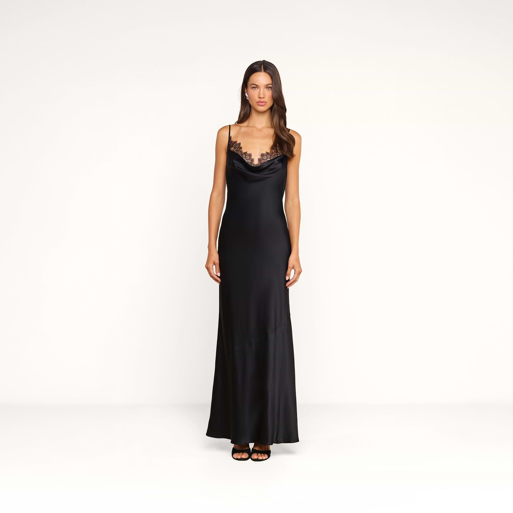 A woman stands in the Selene Silk Gown, a black evening dress with lace trim and thin straps, paired with black heels, against a plain white background.