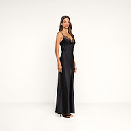 A woman stands in a minimal white studio, facing slightly to the side, wearing the Selene Silk Gown—a black evening dress featuring lace detail on the neckline—paired with black high heels and her long straight hair cascading down.