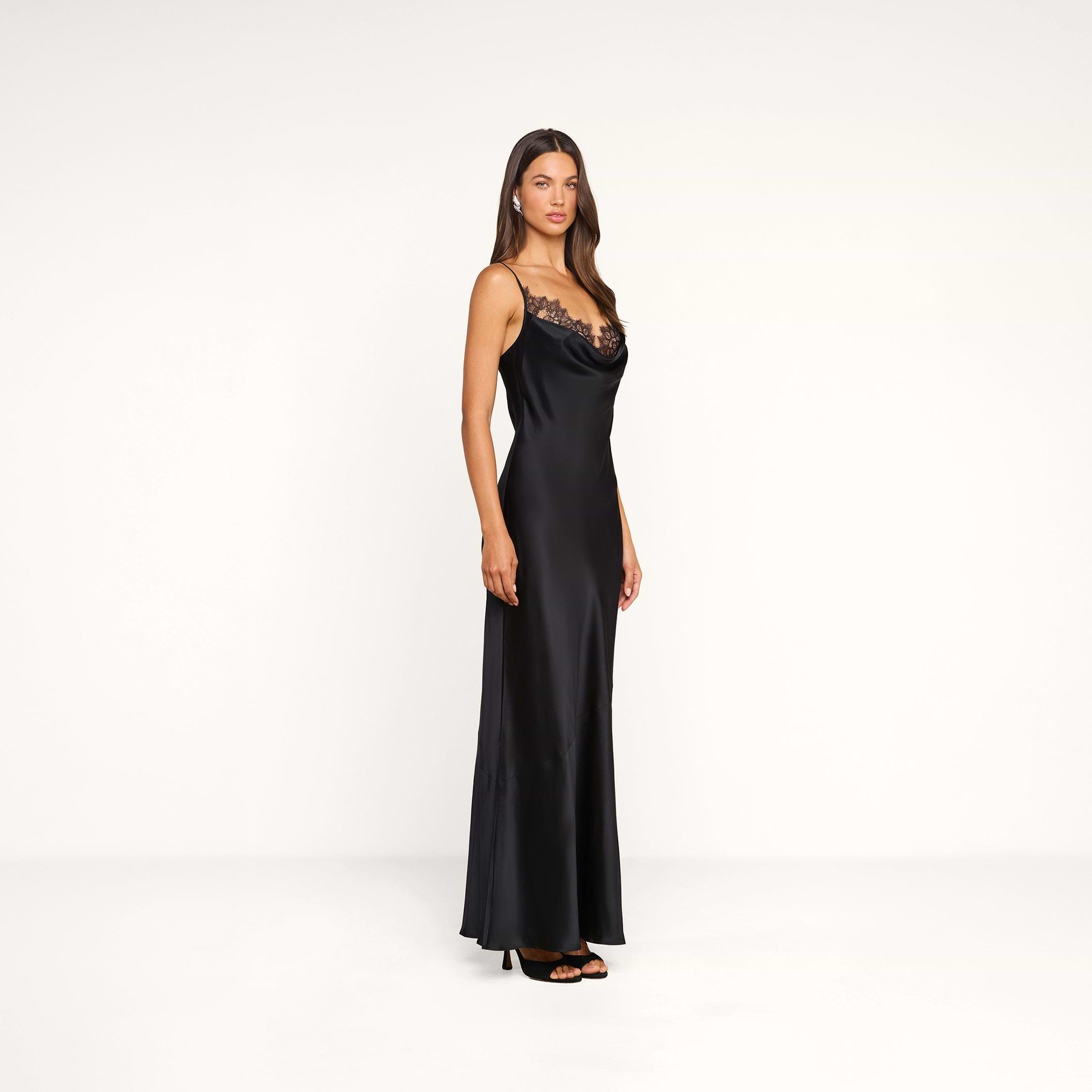 A woman stands in a minimal white studio, facing slightly to the side, wearing the Selene Silk Gown—a black evening dress featuring lace detail on the neckline—paired with black high heels and her long straight hair cascading down.