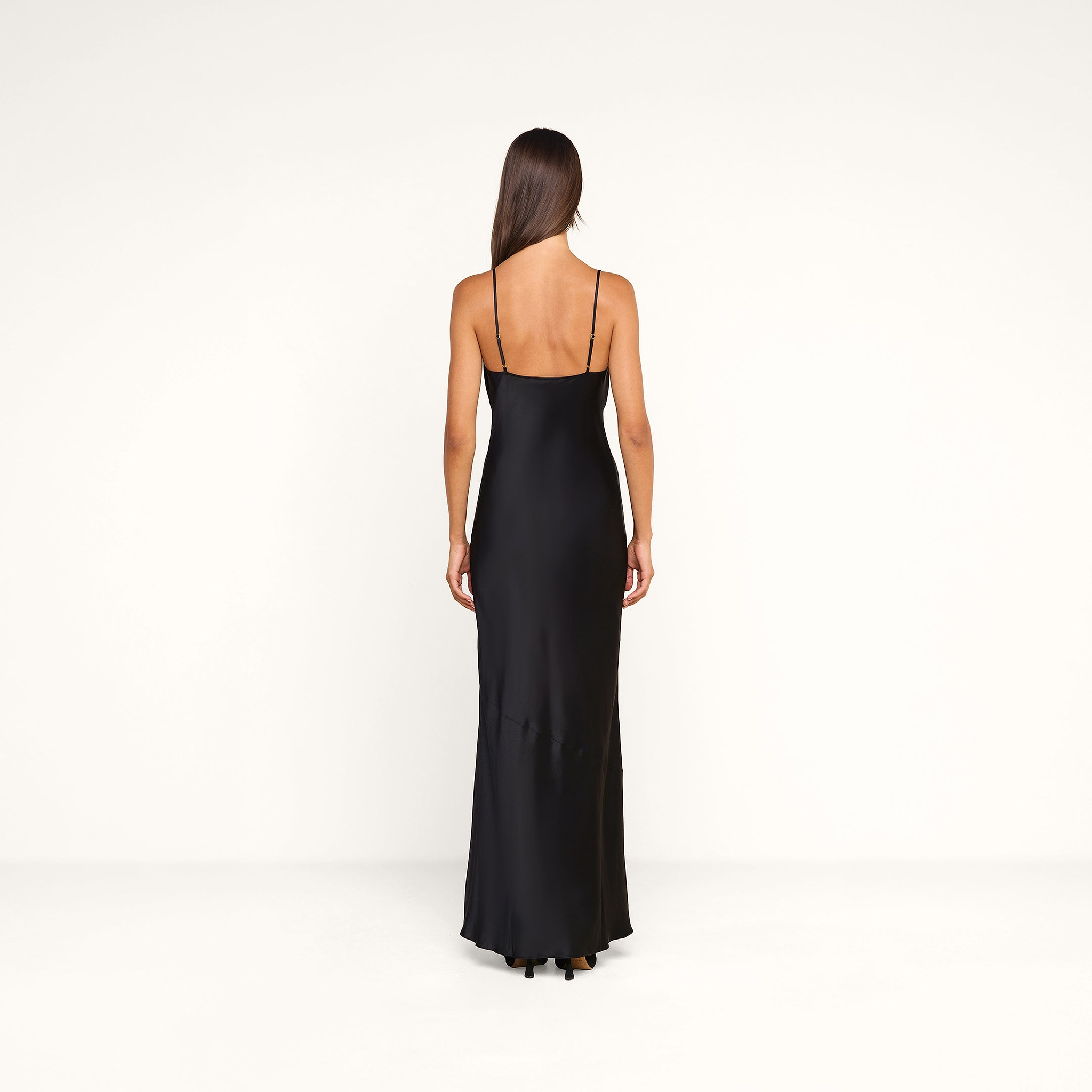 A woman with long brown hair stands facing away, wearing the Selene Silk Gown—a black evening dress with thin straps—in a simple white studio setting.