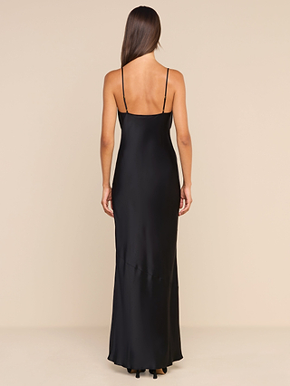 A woman with long brown hair models the Selene Silk Gown—a black, floor-length evening dress with thin straps—seen from the back as she stands against a beige background.