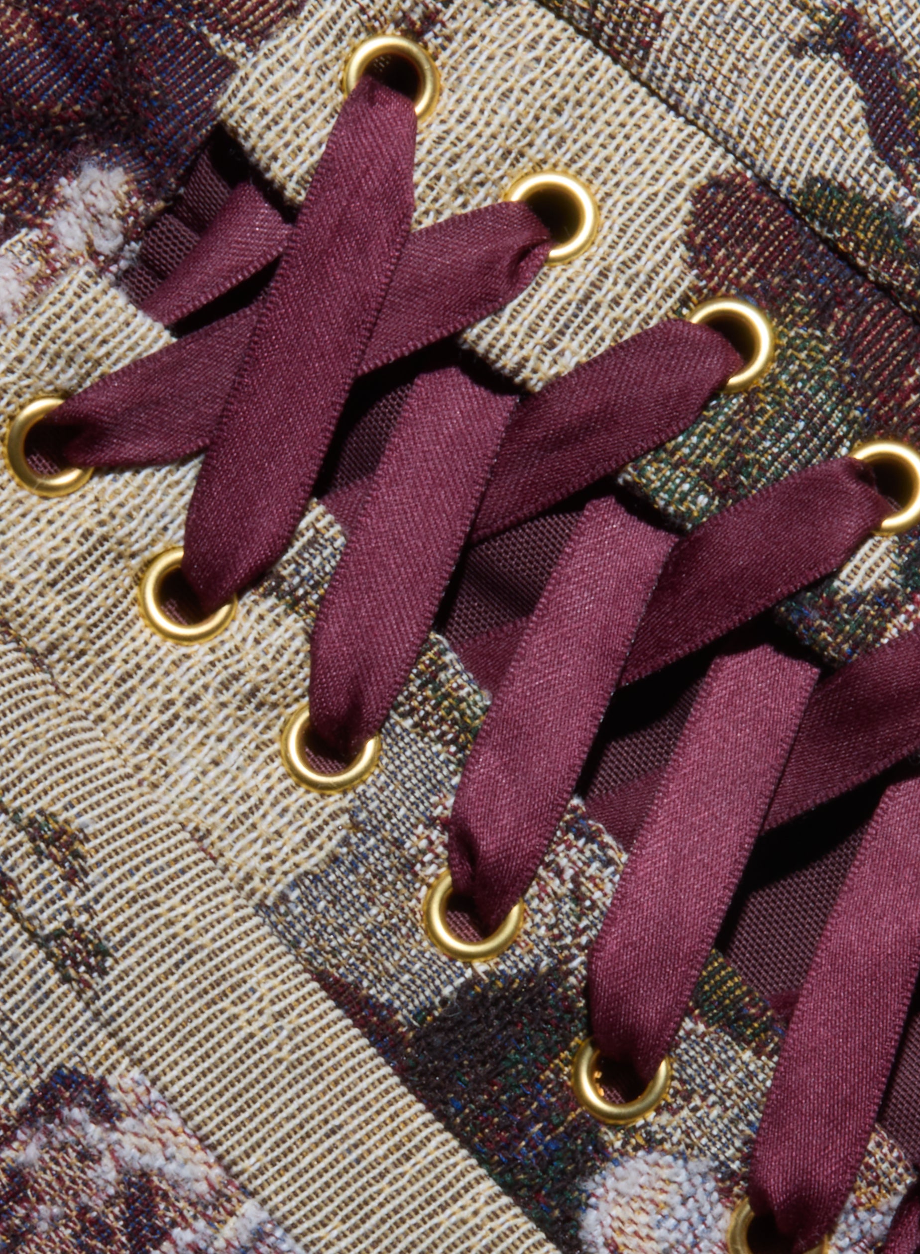 Close-up of maroon shoelaces crisscrossed through gold eyelets on a textured, patterned fabric background, evoking the corset-inspired back detail of the Renee Tapestry Mini Dress.