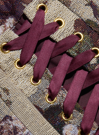 Close-up of maroon shoelaces threaded through gold eyelets on a textured, patterned fabric, part of the Renee Tapestry Mini Dress.