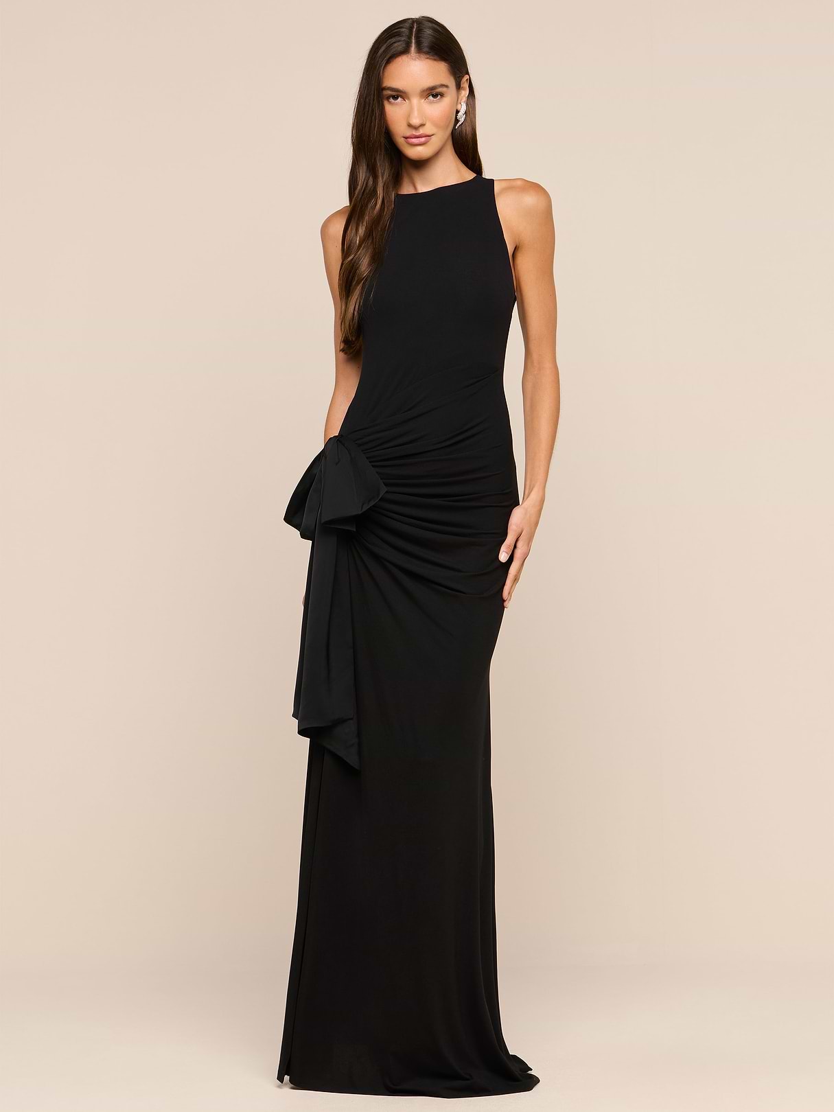 A woman poses against a crisp white backdrop in the Frost Gown, a full-length black dress featuring a draped hip detail. Her loose waves cascade over one shoulder, capturing sophisticated New York Fashion Week-inspired style.