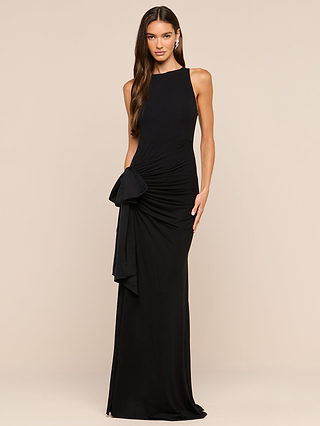 A woman in the Frost Gown—a sleeveless, full-length black dress featuring a striking satin side bow detail—stands against a plain beige background, looking at the camera with one hand by her side and the other resting on her hip.