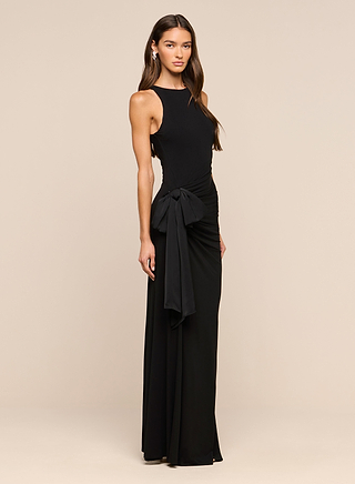 A woman wears the full-length, sleeveless Frost Gown in black, featuring a striking satin side bow detail. She stands against a plain beige background with long, wavy hair, posing with one hand resting by her side.