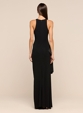 A woman is seen from the back wearing the Frost Gown, a long, sleeveless black dress with a high neckline. This full-length gown features a fitted silhouette and subtle ruffle detailing on one side. The background is plain and beige.