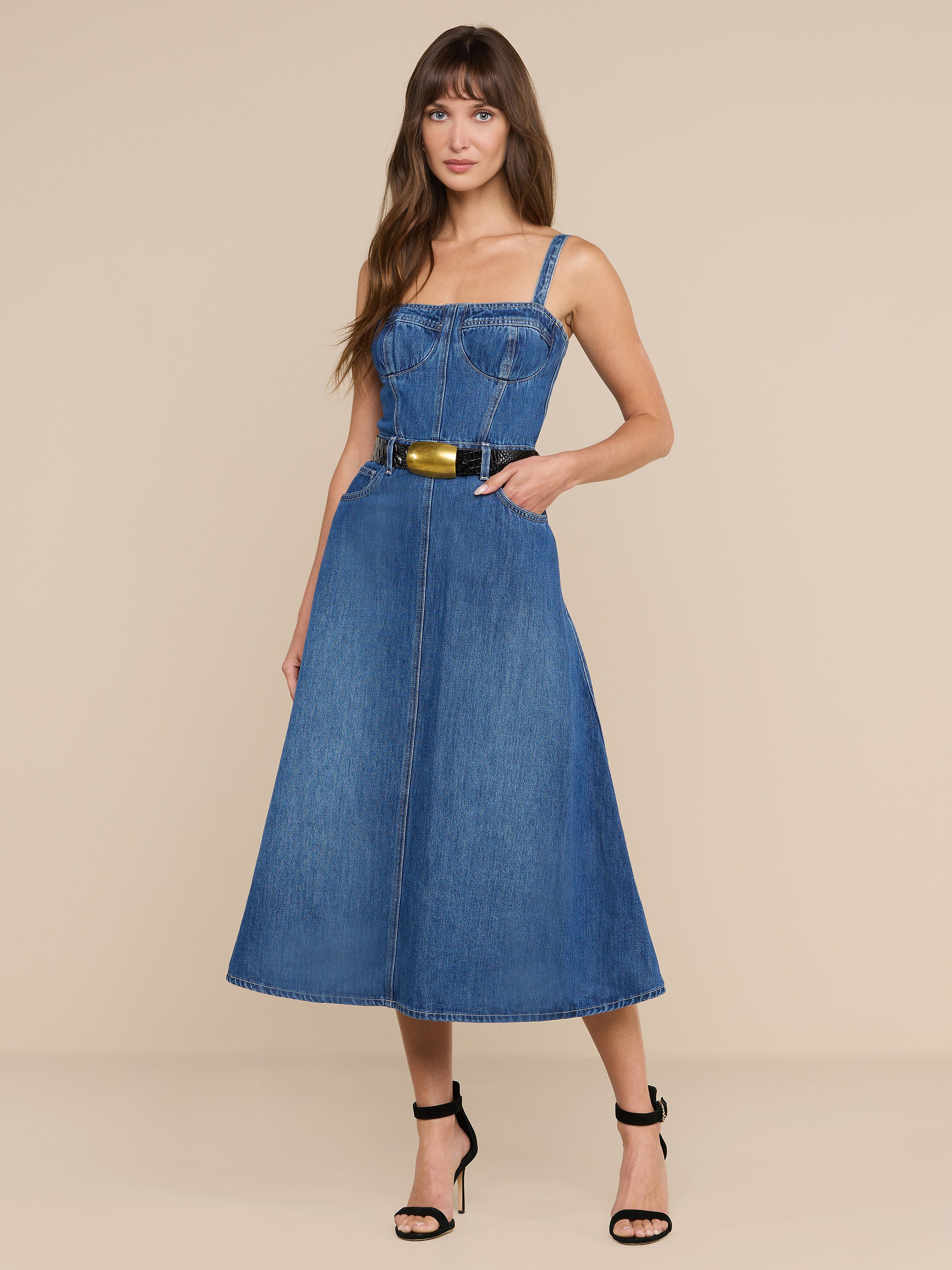 Una donna posa su uno sfondo beige indossando labito bustier Jovie: un modello in denim senza maniche con corpetto a corsetto, cintura in vita e gonna a trapezio. Lo abbina a tacchi neri, con i lunghi capelli castani sciolti e una mano in tasca.