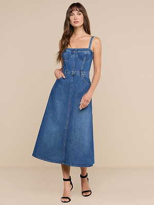 Su uno sfondo beige, una donna dai lunghi capelli castani indossa il Jovie Bustier Dress — un abito senza maniche in denim blu con corpetto a corsetto e gonna a trapezio — abbinato a sandali neri con tacco alto, con una mano infilata con disinvoltura in tasca.