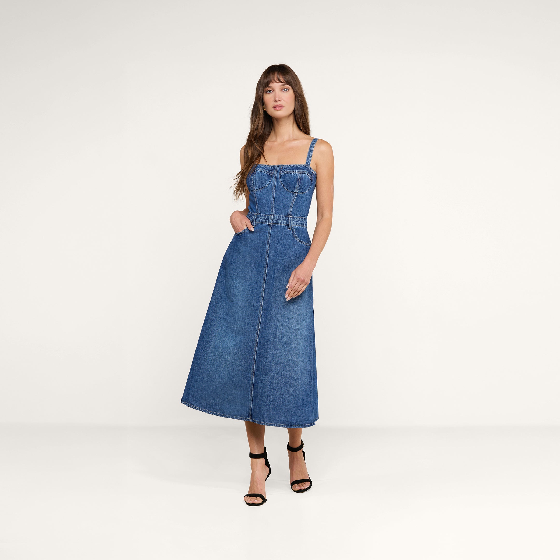 Su uno sfondo beige, una donna dai lunghi capelli castani indossa il Jovie Bustier Dress — un abito senza maniche in denim blu con corpetto a corsetto e gonna a trapezio — abbinato a sandali neri con tacco alto, con una mano infilata con disinvoltura in tasca.
