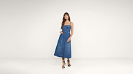 A woman stands against a plain background wearing a sleeveless, belted denim midi dress and black high-heeled sandals.