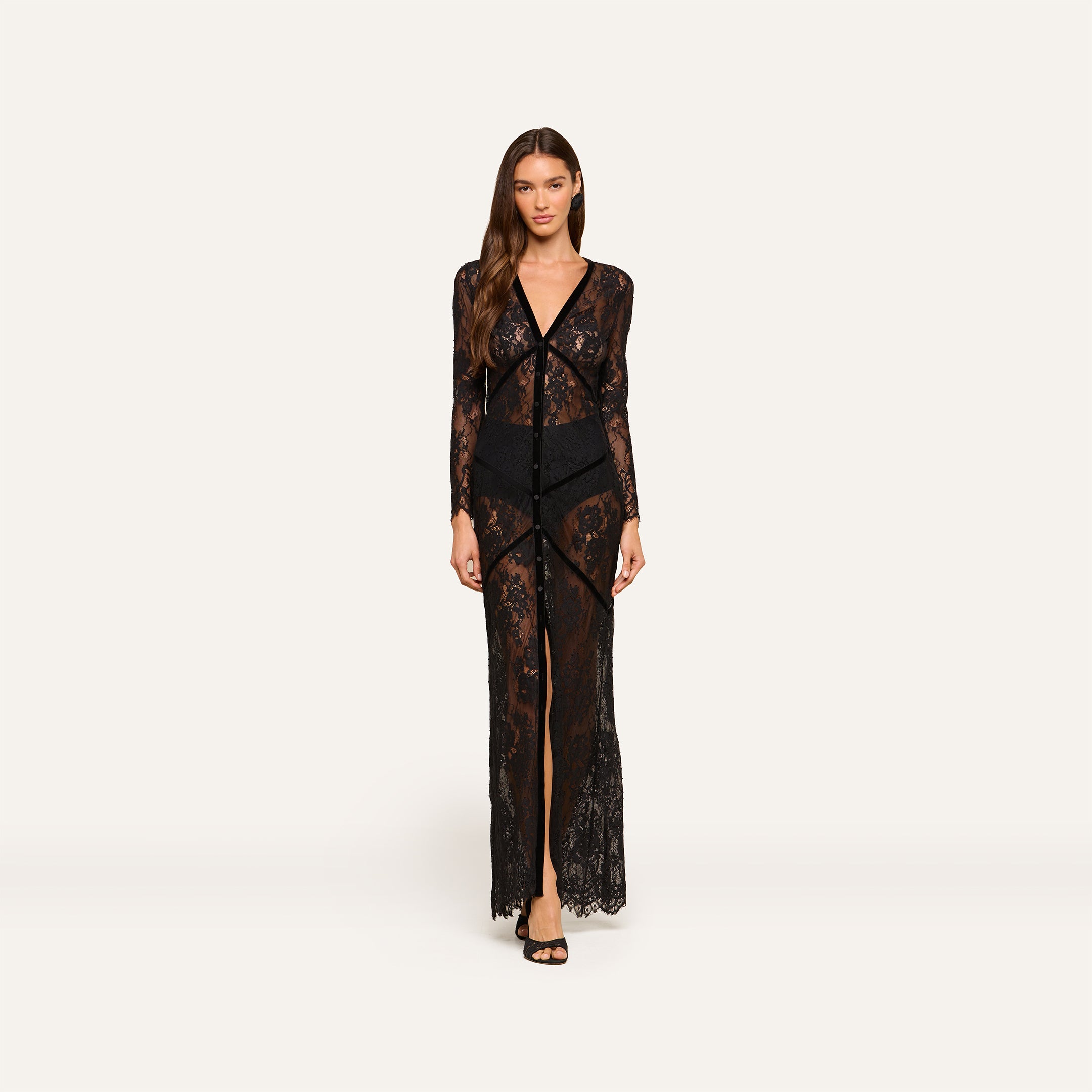 A woman stands facing forward, wearing the Poesy Lace Maxi Dress— a black lace v-neck maxi with long sleeves and black undergarments. The sheer dress is set against a plain light background.