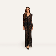 A woman stands facing forward, wearing the Poesy Lace Maxi Dress— a black lace v-neck maxi with long sleeves and black undergarments. The sheer dress is set against a plain light background.