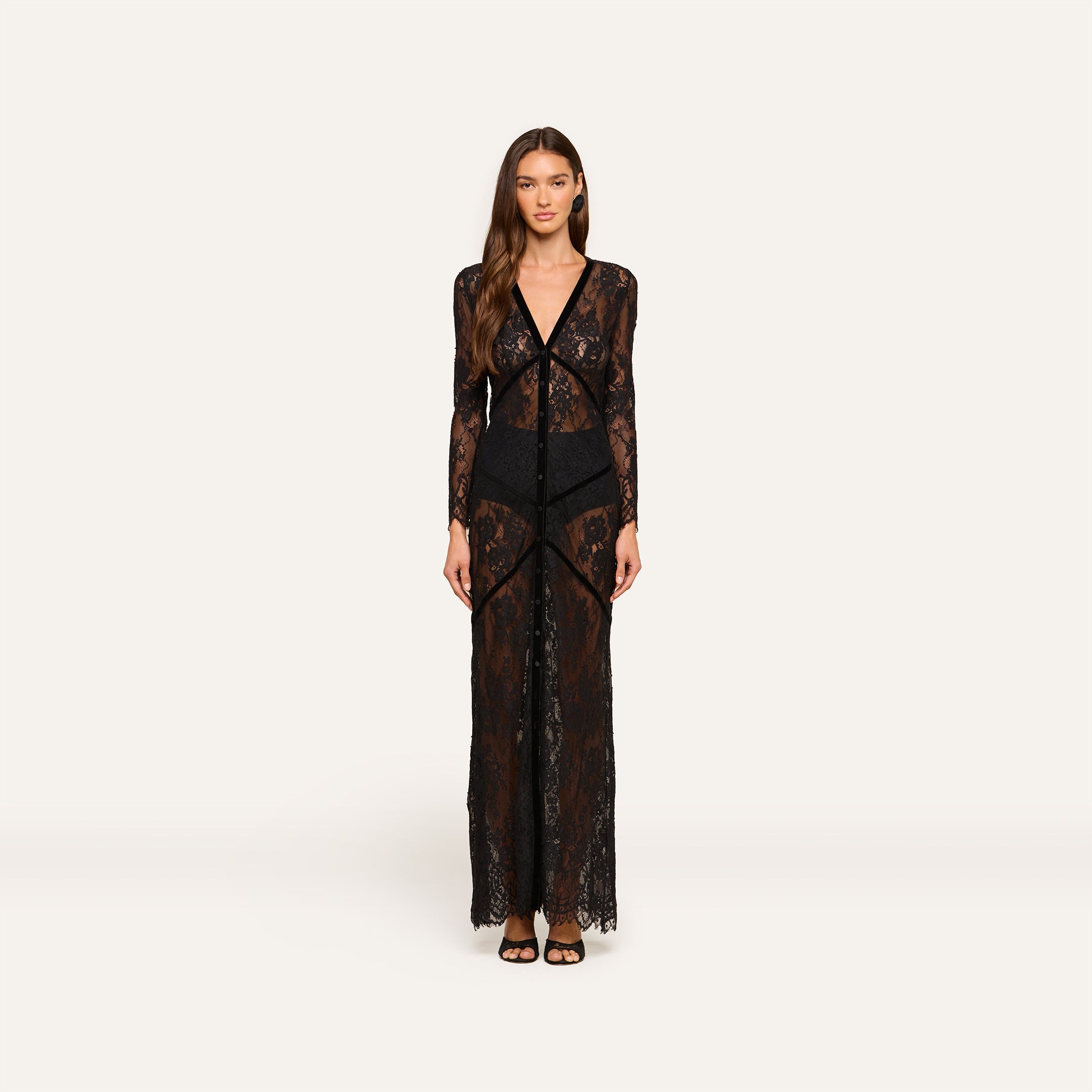 A woman faces forward wearing the Poesy Lace Maxi Dress—a black lace, ankle-length maxi featuring sheer long sleeves and black undergarments. She has long brown hair, a neutral expression, and is styled with black sandals against a plain white background.