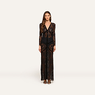 A woman faces forward wearing the Poesy Lace Maxi Dress—a black lace, ankle-length maxi featuring sheer long sleeves and black undergarments. She has long brown hair, a neutral expression, and is styled with black sandals against a plain white background.