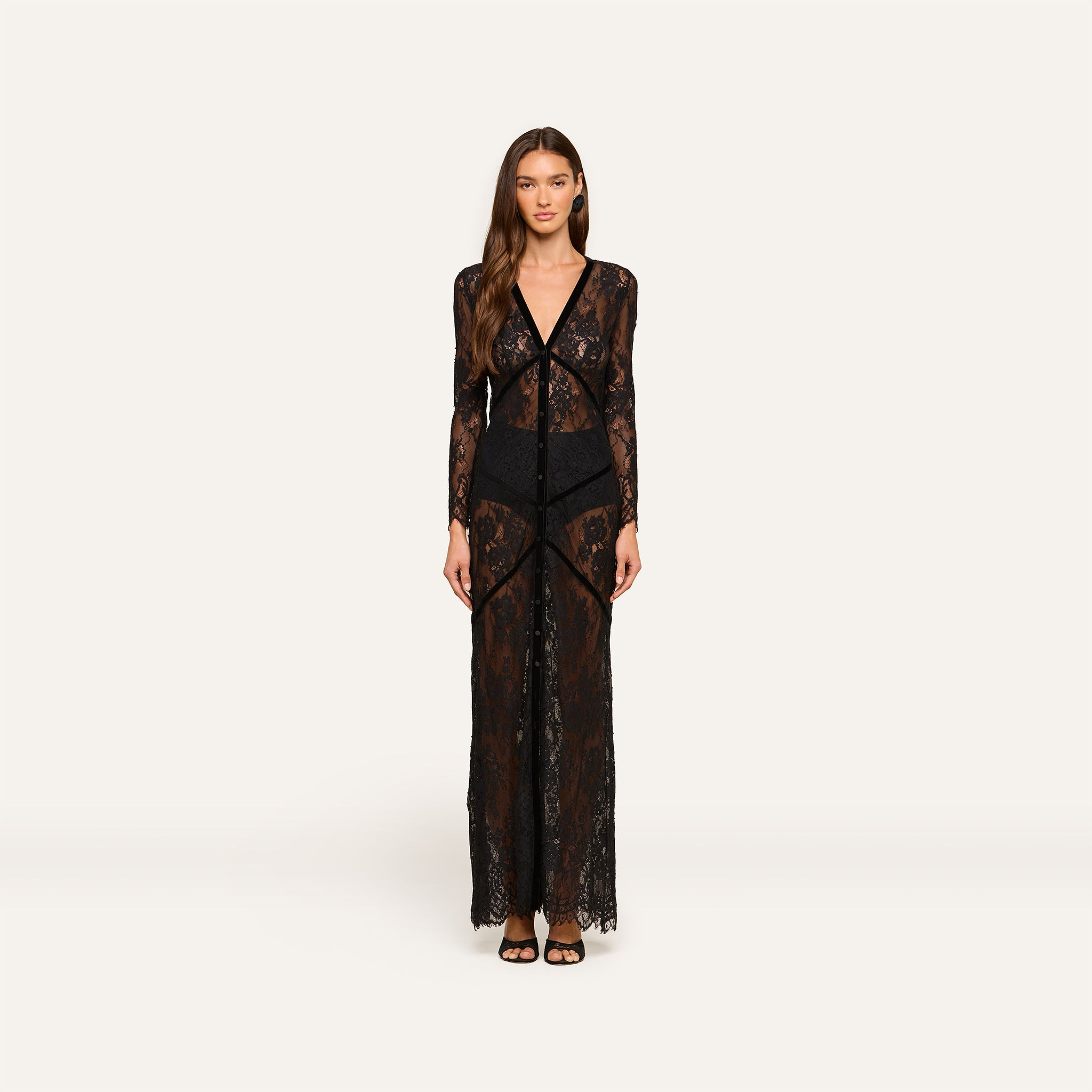A woman faces forward wearing the Poesy Lace Maxi Dress—a black lace, ankle-length maxi featuring sheer long sleeves and black undergarments. She has long brown hair, a neutral expression, and is styled with black sandals against a plain white background.